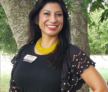 Female Employee standing outdoors in front of a tree in the sunlight wearing a black shirt with yellow necklace