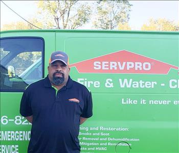 Male Employee of SERVPRO wearing a baseball cap and black short sleeve collared shirt standing in front of green company van